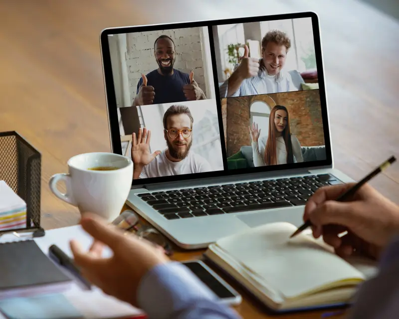 A person holds a coffee mug and takes notes in an open notebook while viewing a four-way video call on a laptop. The screen shows four smiling participants — two giving thumbs up and two waving — suggesting an upbeat remote group interview or team meeting taking place from a home office desk.