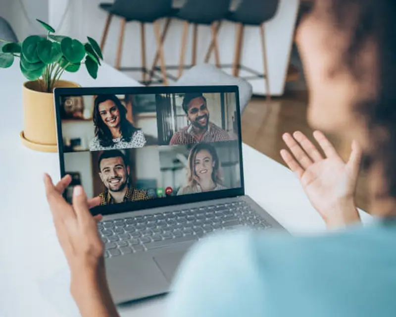 A person seen from behind waves toward a laptop screen showing a four-way video call with three smiling participants. A potted plant sits nearby in a bright living space. The scene captures the warmth of a remote interview or virtual team meeting conducted from a home environment.