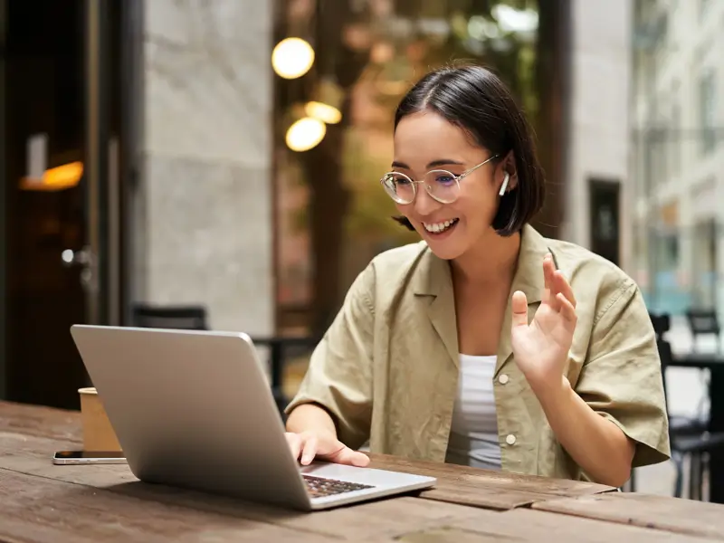 Woman with glasses and earbuds smiling and waving at a laptop screen at an outdoor cafe table