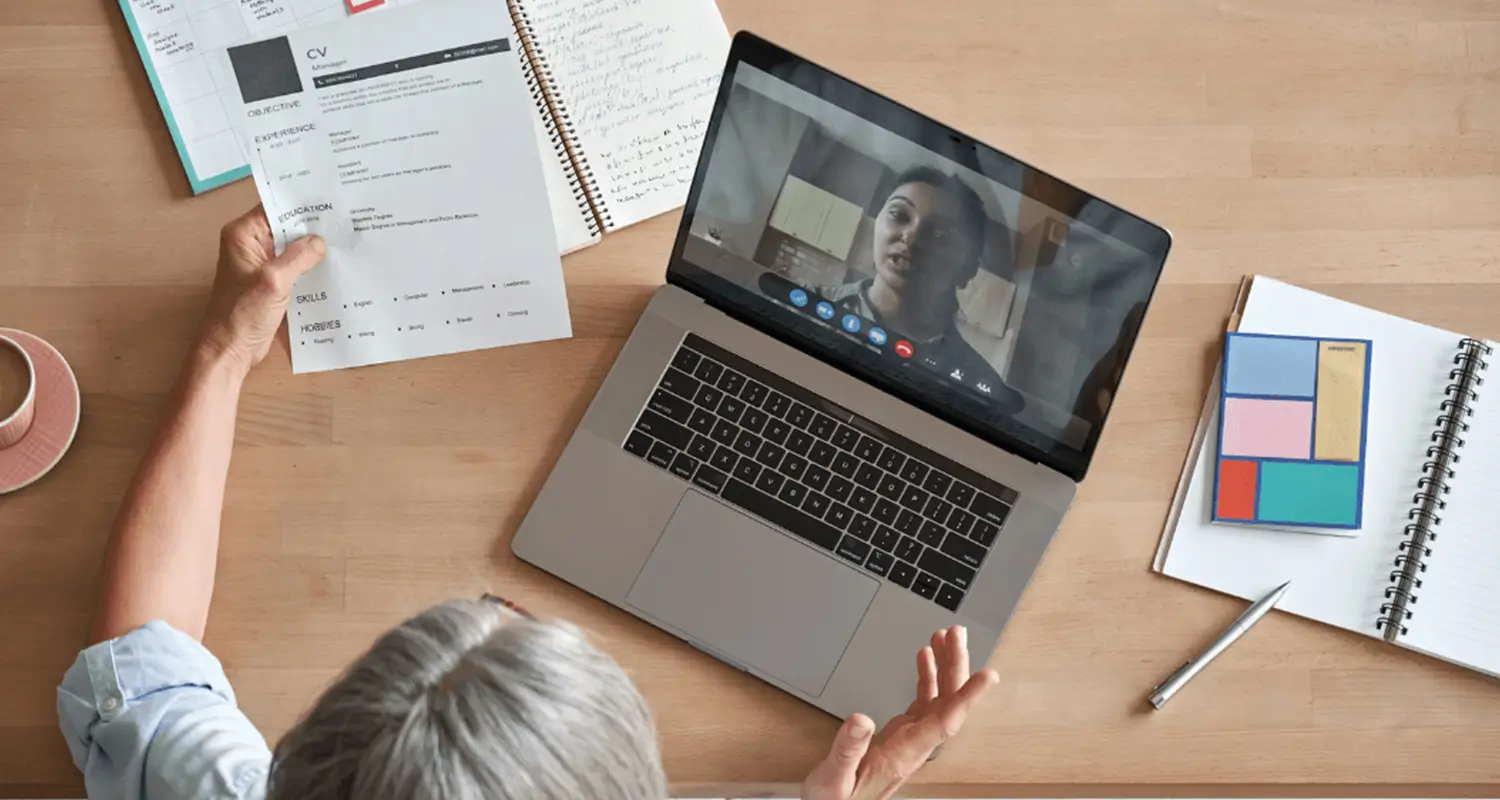 Overhead view of a hiring manager holding a printed CV while conducting a video interview on a laptop. A second participant is visible on screen. Notebooks, colorful sticky notes, a pen, and a mug are spread across the desk.