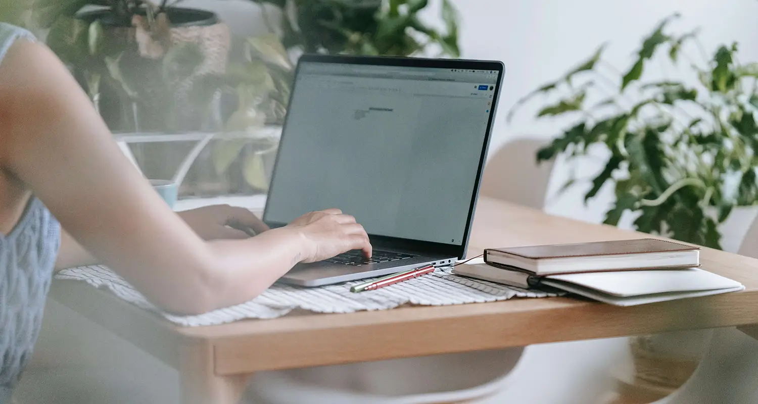 A person types on a laptop at a light wood desk in a bright, plant-filled home workspace. Notebooks and a pencil are arranged beside the laptop, suggesting a focused remote work setup.