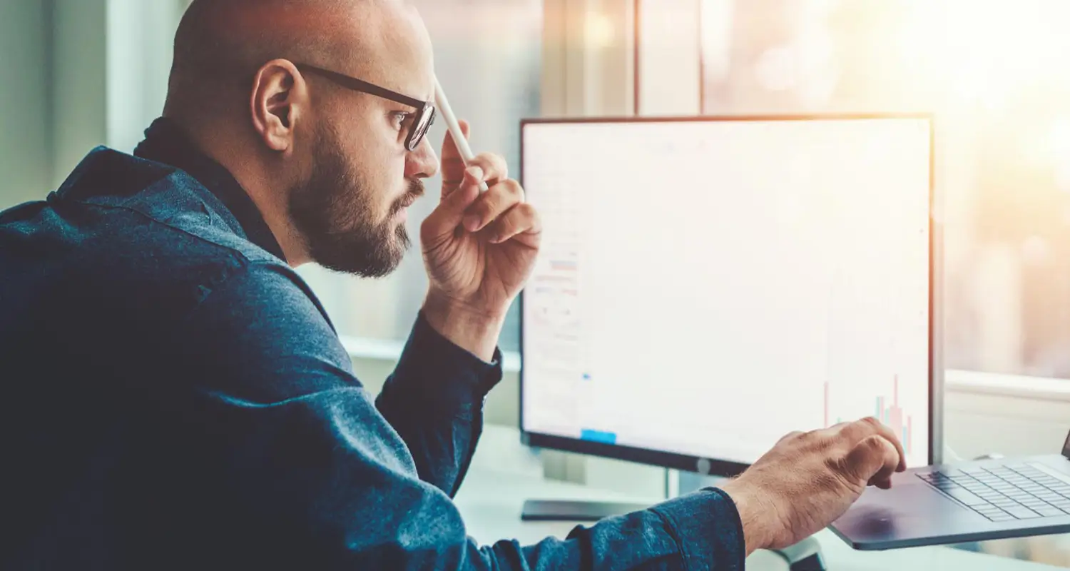 A bald man wearing glasses and a dark hoodie sits at a dual-monitor workstation, holding a pen to his temple in a thoughtful pose while reviewing data on screen. Charts and graphs are faintly visible on the monitors, suggesting he is analyzing remote team performance metrics in a professional office setting.