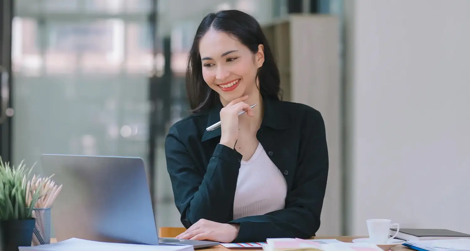 A smiling woman in a dark blazer rests her chin on her hand while holding a pen and reviewing documents on her laptop in a modern office. Papers, a coffee cup, and a plant are visible on her desk, suggesting a manager thoughtfully preparing for or conducting a remote employee performance review.