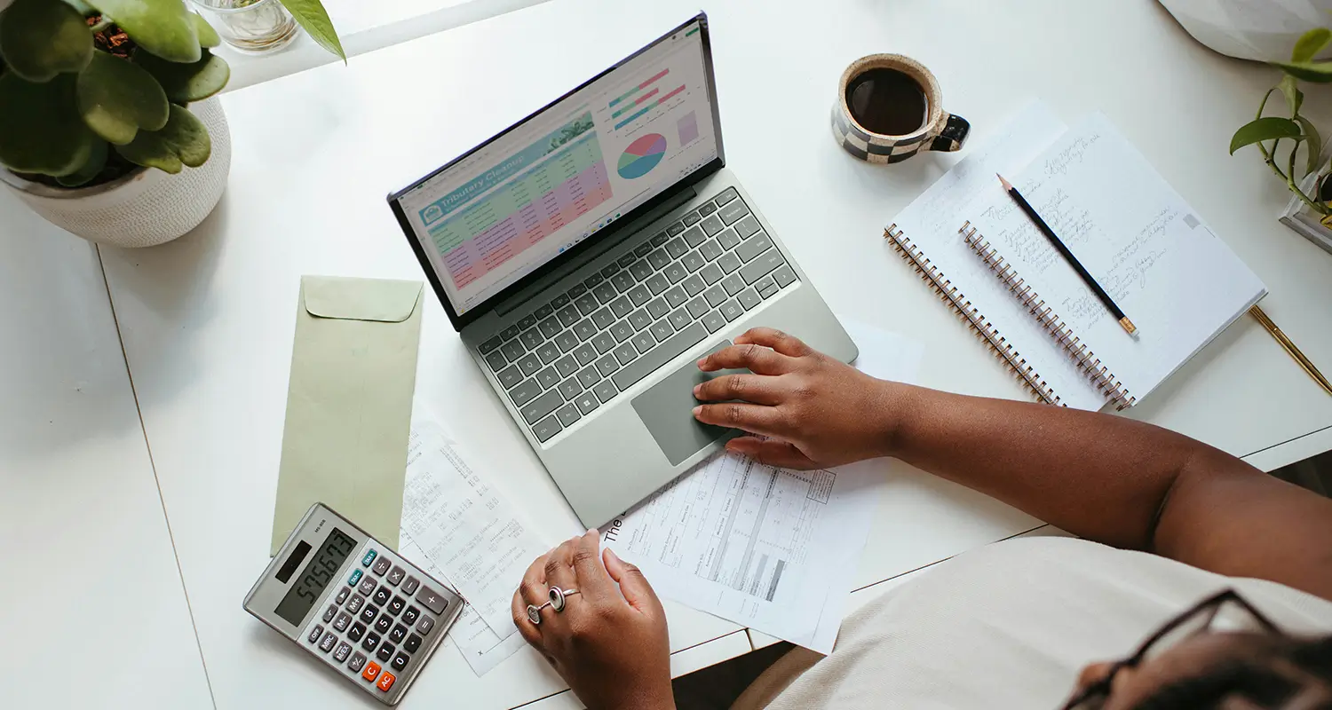 Person working at a desk with a laptop displaying a colorful spreadsheet and charts, alongside a calculator, notebook, and coffee mug.