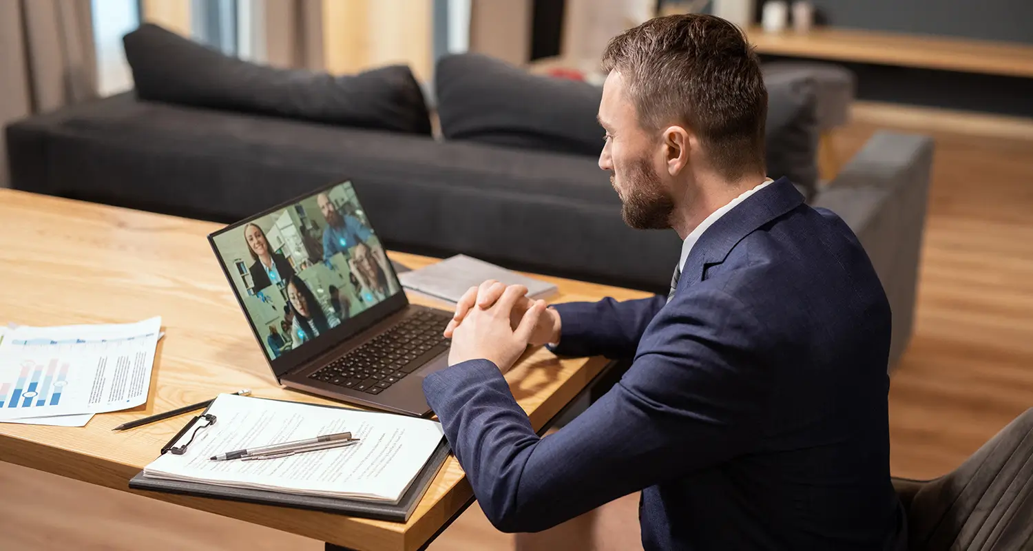Business professional in a navy suit joining a video conference with four remote colleagues displayed on a laptop, notes and charts on the desk.