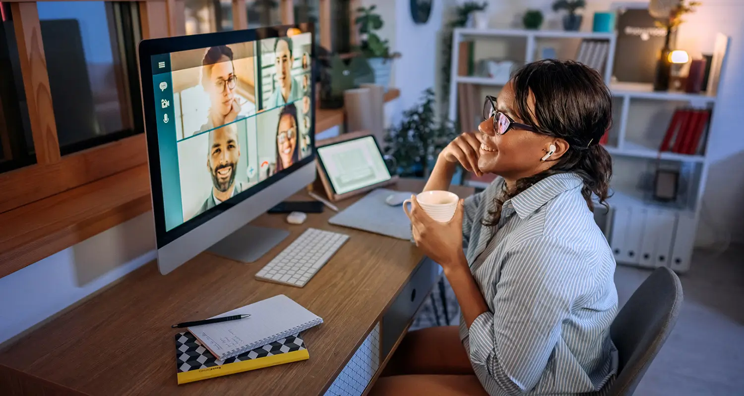 A smiling woman with glasses and wireless earbuds holds a coffee mug while attending a video call on a large desktop monitor showing six remote colleagues in a grid layout, seated at a wooden home office desk with a notebook, tablet, and shelving unit in the background.