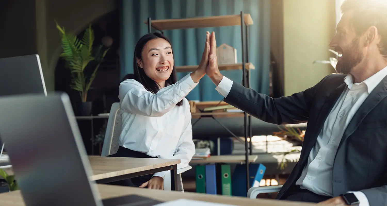 Smiling woman high-fiving a male colleague at an office desk, celebrating a team achievement with a laptop open nearby