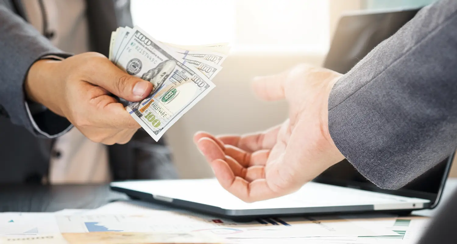A hand passing US dollar bills to another person's open palm over a desk with financial charts, representing incentive bonus payment