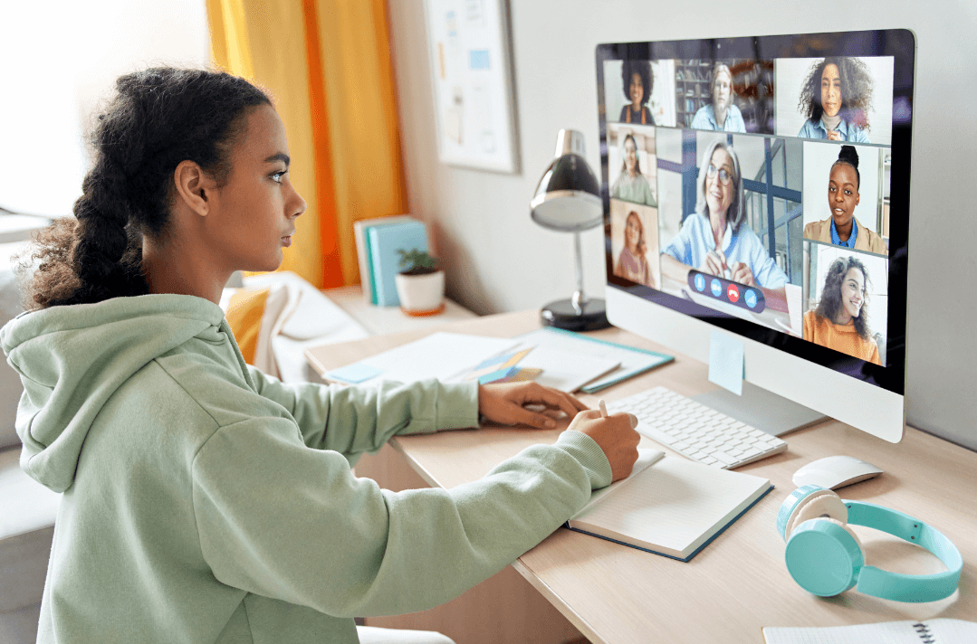 A young woman with curly hair sits at a home desk at night, looking attentively at a large desktop monitor displaying a video call with a woman in a colorful setting. Headphones are nearby, depicting a remote employer conducting a video interview or check-in session.
