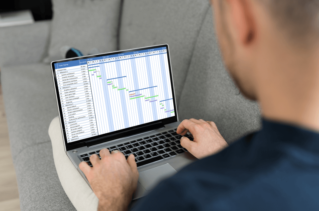 A man viewed from behind sits on a couch reviewing a Gantt chart project management timeline on a laptop screen. The colorful chart shows multiple tasks and milestones across January and February, illustrating the use of project tracking tools for remote employee productivity.
