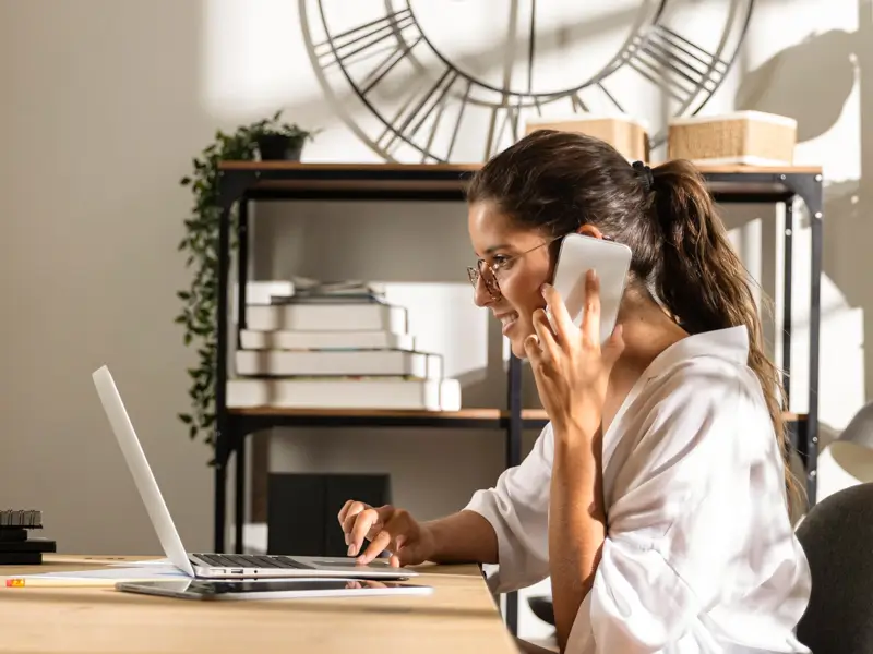Woman with glasses talking on phone while typing on a laptop at a home office desk with shelving behind her