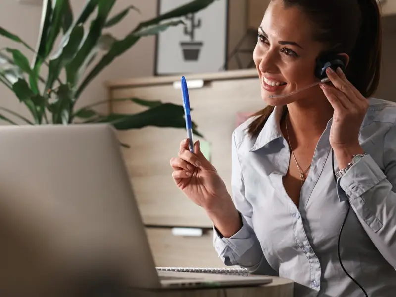 Woman with a headset smiling and holding a pen while looking at a laptop in a home office setting