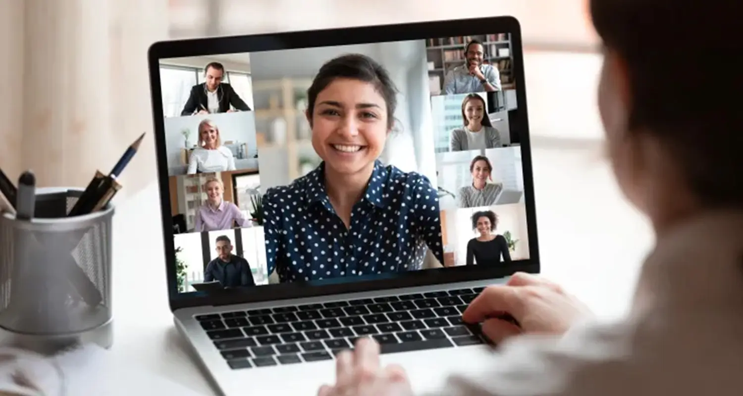 A manager seen from behind participates in a video call on a laptop, with eight remote team members displayed in a grid layout on screen. A pen holder sits on the desk beside the laptop, illustrating a leader conducting an all-hands or team alignment meeting with distributed employees.