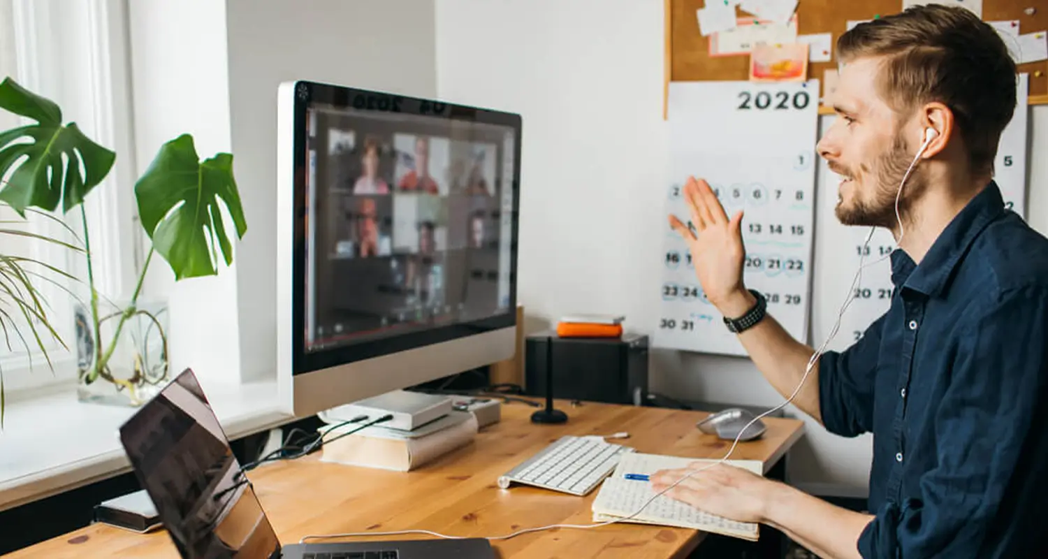 A smiling man with earbuds waves at a desktop iMac displaying a video call with multiple participants, while sitting at a wooden home office desk. A wall calendar, a cork board, a laptop, and a large monstera plant are visible in the background, depicting an engaged remote worker in a virtual team meeting.