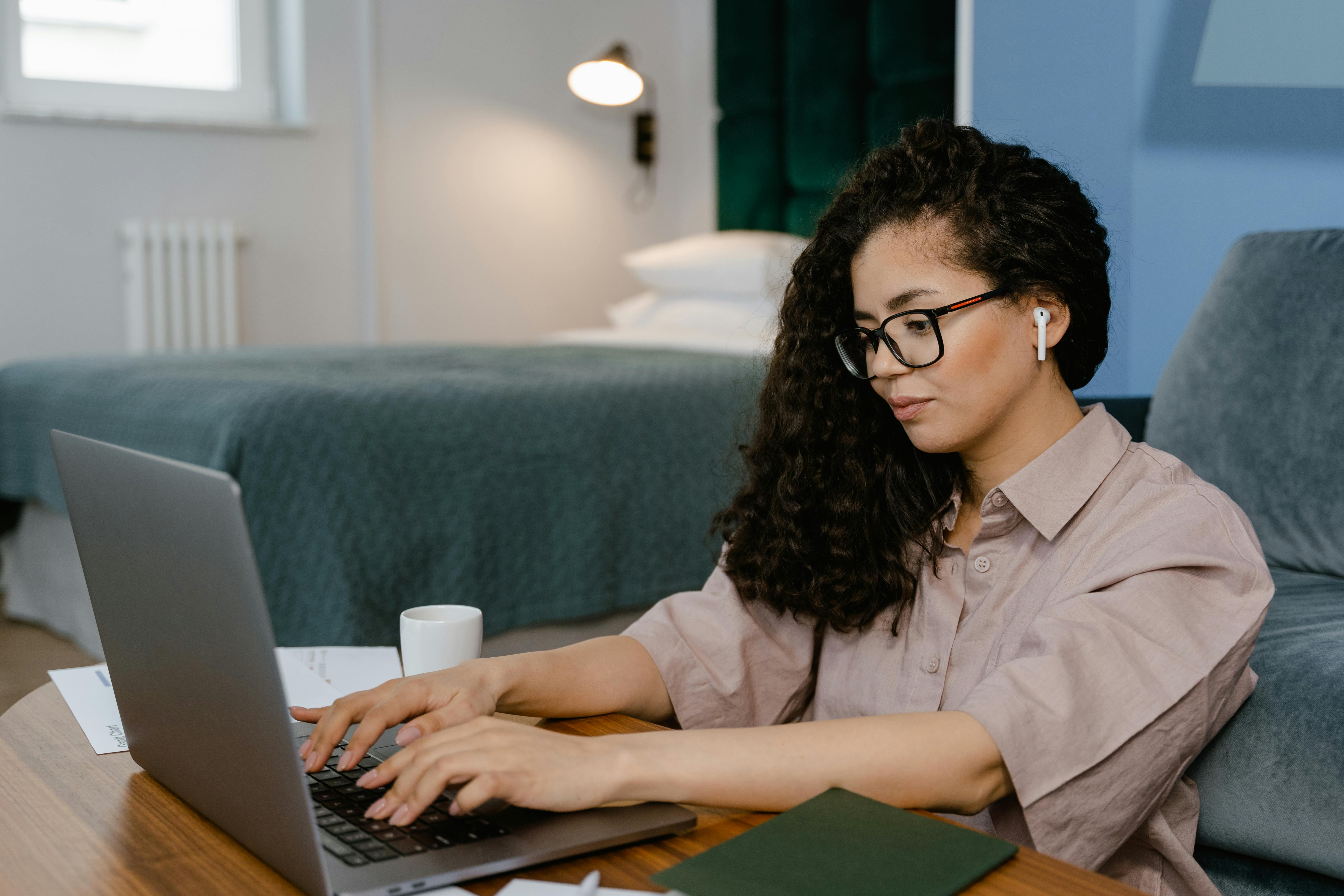 A woman with curly hair, glasses, and a wireless AirPod types on a laptop at a low wooden table, with a white mug and scattered papers nearby. A bed and blue-tinted wall in the background suggest a hotel room or home workspace used for remote sales work.