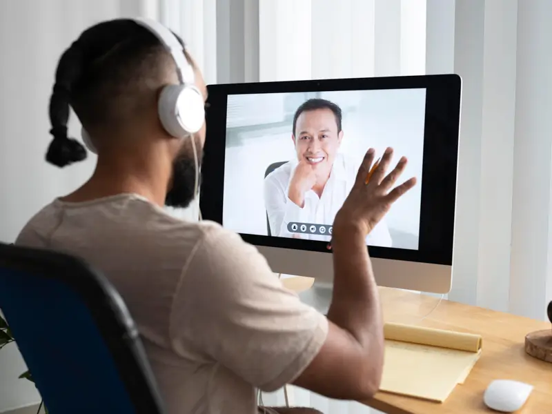 Man with headphones waving at a smiling candidate on a desktop monitor during a one-on-one video interview