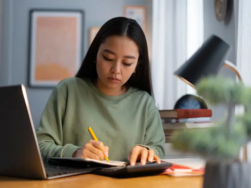 Young woman in a green sweatshirt writing notes with a pencil next to a laptop at a home desk with a lamp