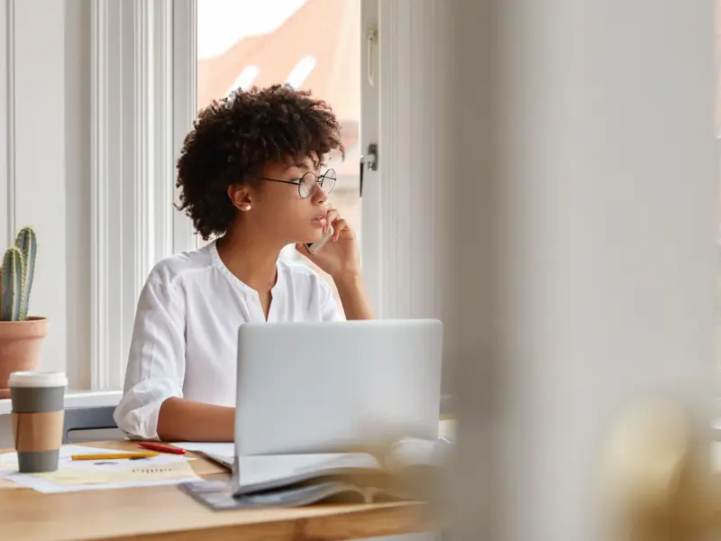 Woman with glasses and natural hair resting her chin on her hand while looking out a window beside a laptop