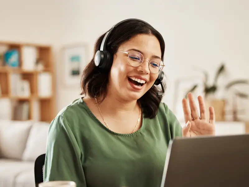 Woman wearing headphones and glasses smiling and waving at a laptop screen during a remote video call