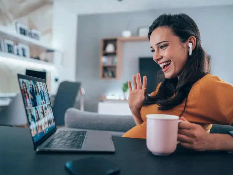 Woman with wireless earbuds smiling and waving during a video call on her laptop from a home office