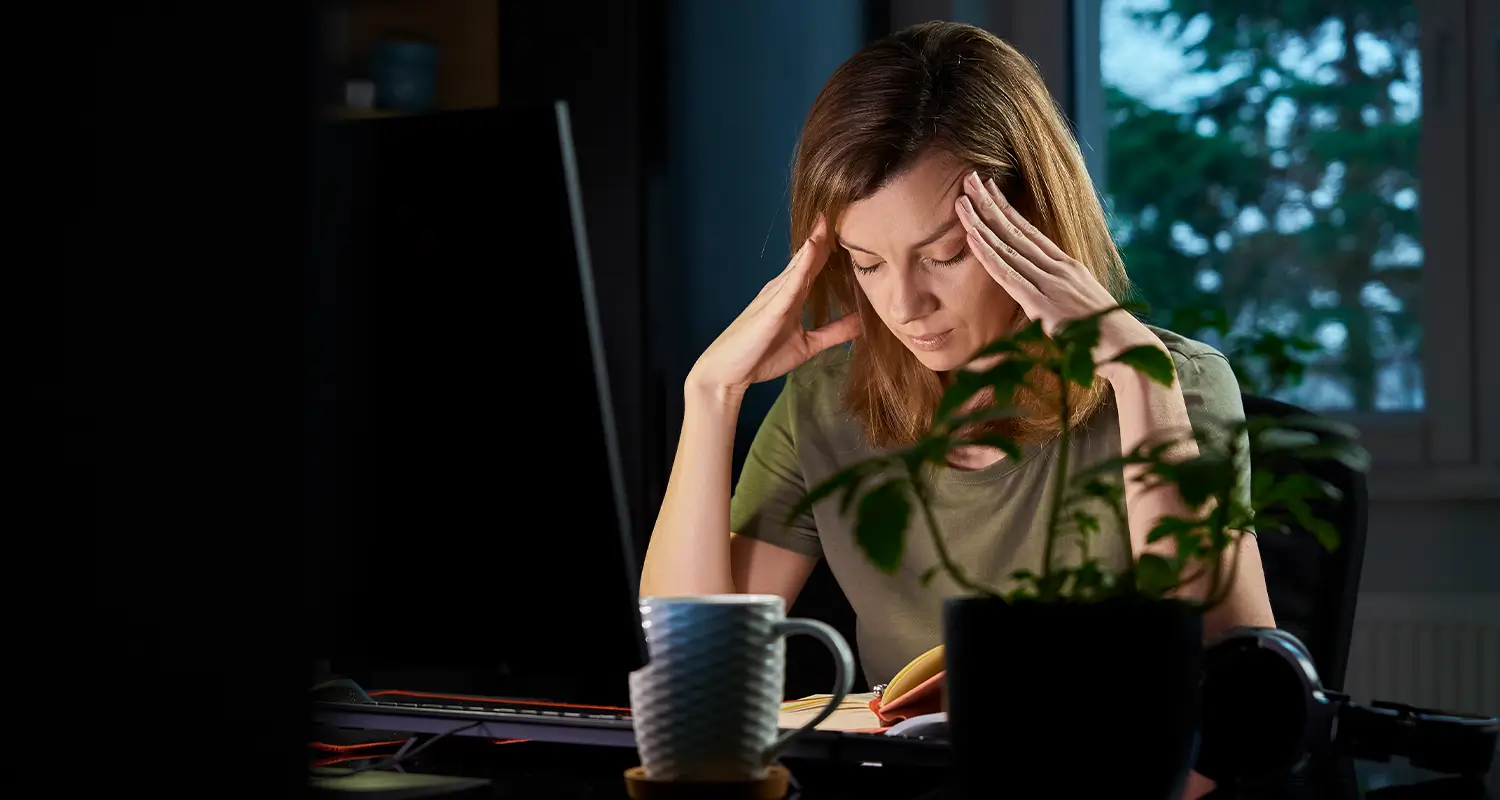A woman in a green t-shirt holds both hands to her temples with a stressed expression while sitting at her computer late at night. A coffee mug, potted plant, and headphones rest nearby on the dark desk, illustrating the mental strain and burnout risk faced by remote workers who struggle to disconnect.