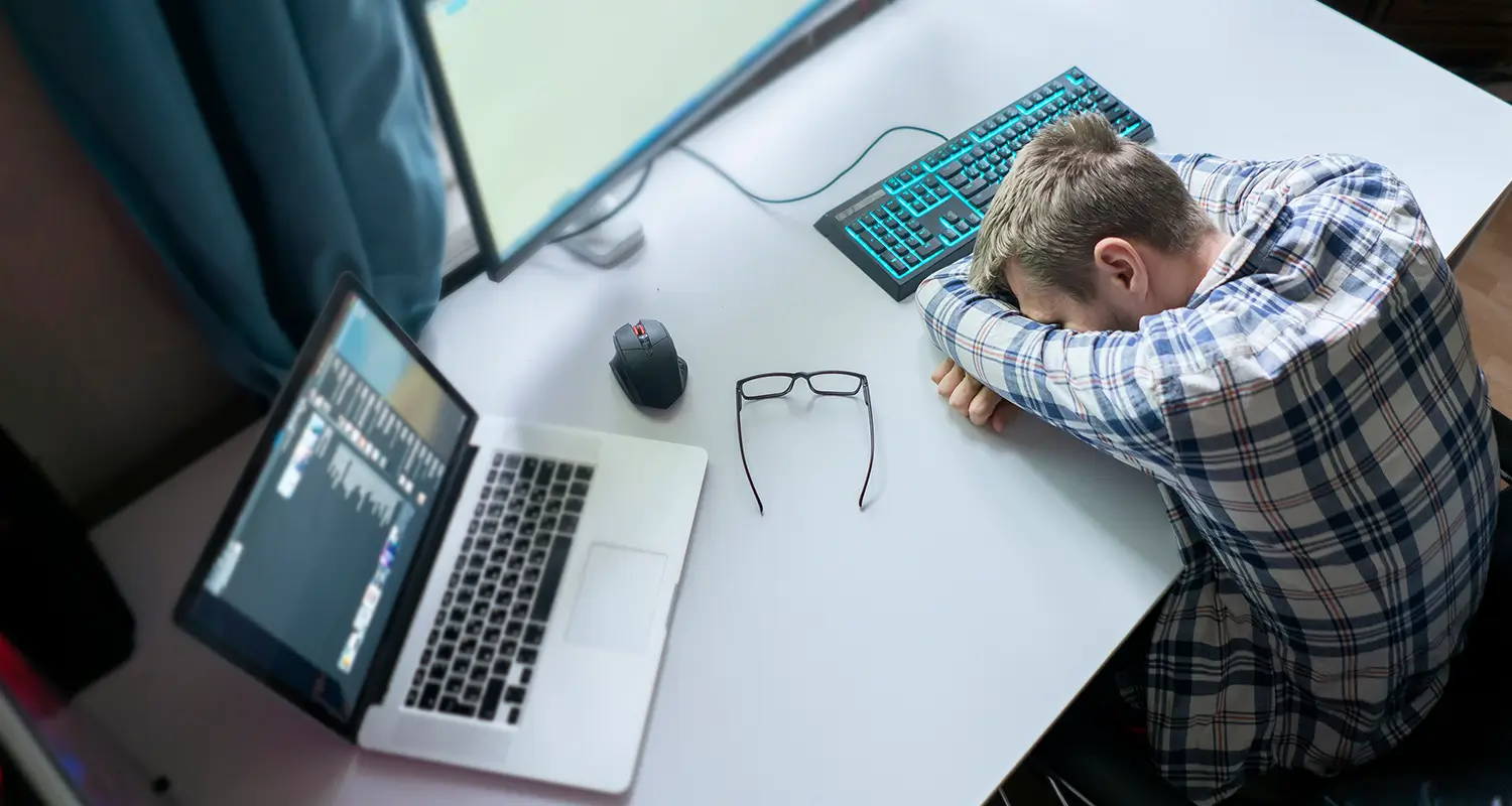 A man in a plaid shirt has collapsed face-down on his desk with his arms folded under his head, his glasses set to the side. A laptop, mouse, and mechanical keyboard are visible on the white desk, conveying extreme exhaustion and burnout from overwork in a remote work environment.