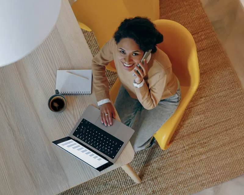 Overhead view of a smiling woman with natural hair wearing a camel-colored top, sitting in a bright yellow chair and talking on a smartphone with an open laptop on her lap. A notebook, pencil, and cup of coffee rest on the table beside her, illustrating a recruiter or hiring professional working remotely.