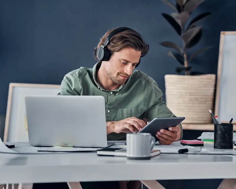 Man wearing over-ear headphones and a green button-up shirt sits at a white desk, using a tablet while an open laptop and coffee mug sit beside him. Sticky notes, pencils, and a plant are visible in the background, depicting a hiring manager or recruiter multitasking while building a candidate pipeline.