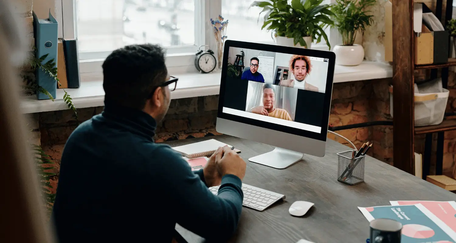 A man with glasses seen from behind sits at a wooden desk in a plant-filled home office, attending a video call on an iMac with three colleagues displayed on screen. A notepad, a pen holder, an Apple keyboard, and printed materials are on the desk, depicting a remote worker staying connected with his distributed team.