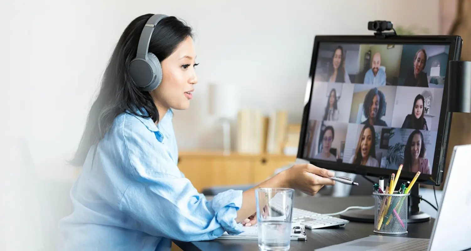 A woman wearing large over-ear headphones sits at a desk and gestures while speaking during a video call, with a monitor displaying a grid of eight remote team members visible on screen. A pen holder, a glass of water, and a webcam are on her desk, illustrating an engaged remote employee actively participating in a virtual team meeting.