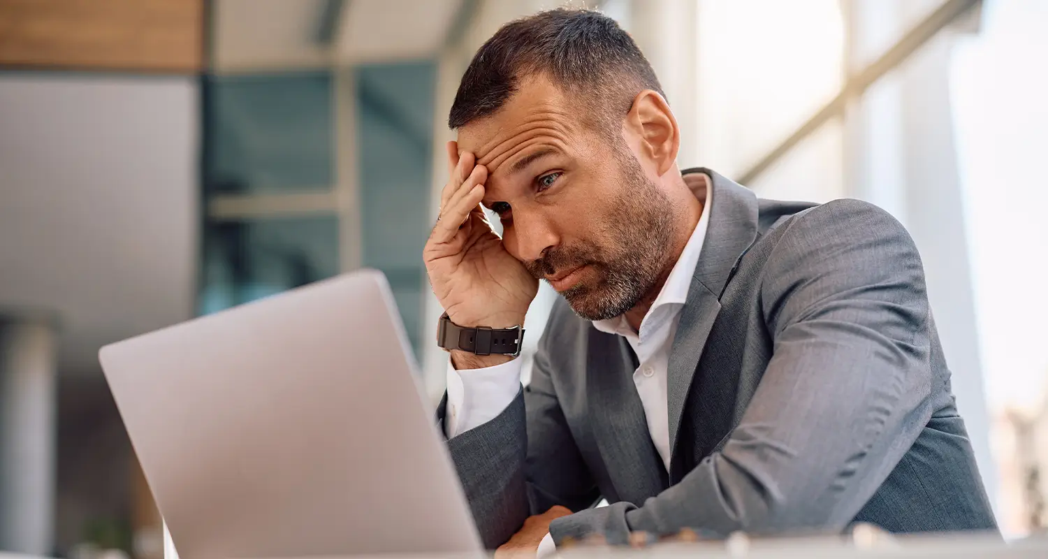 Frustrated businessman in a gray suit leaning his head on his hand while staring at an open laptop, appearing overwhelmed and confused.