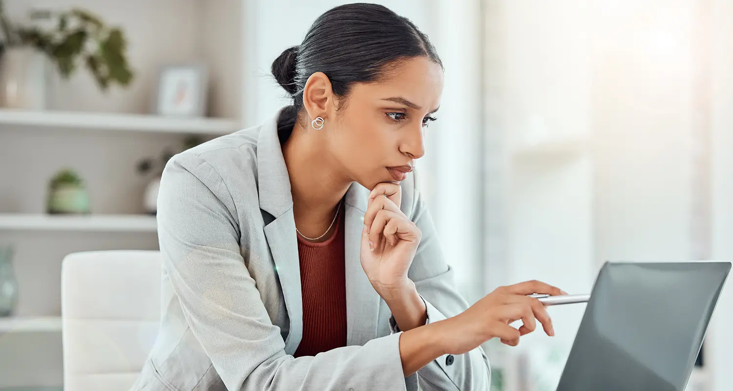 Young professional woman in a blazer thoughtfully pointing at her laptop screen, resting her chin on her hand while working from home.