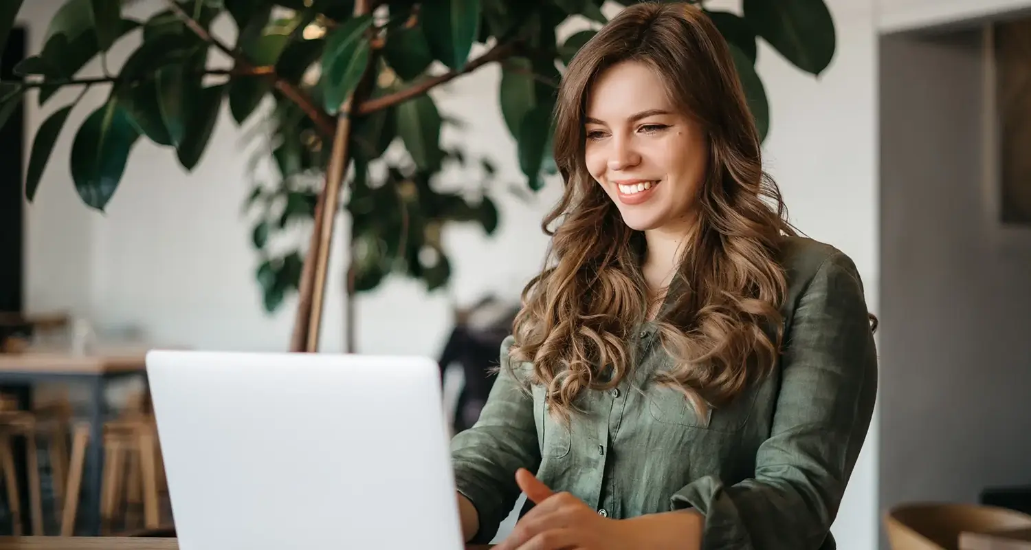 Smiling woman with wavy hair working on a laptop in a bright cafe setting with a large leafy plant in the background.
