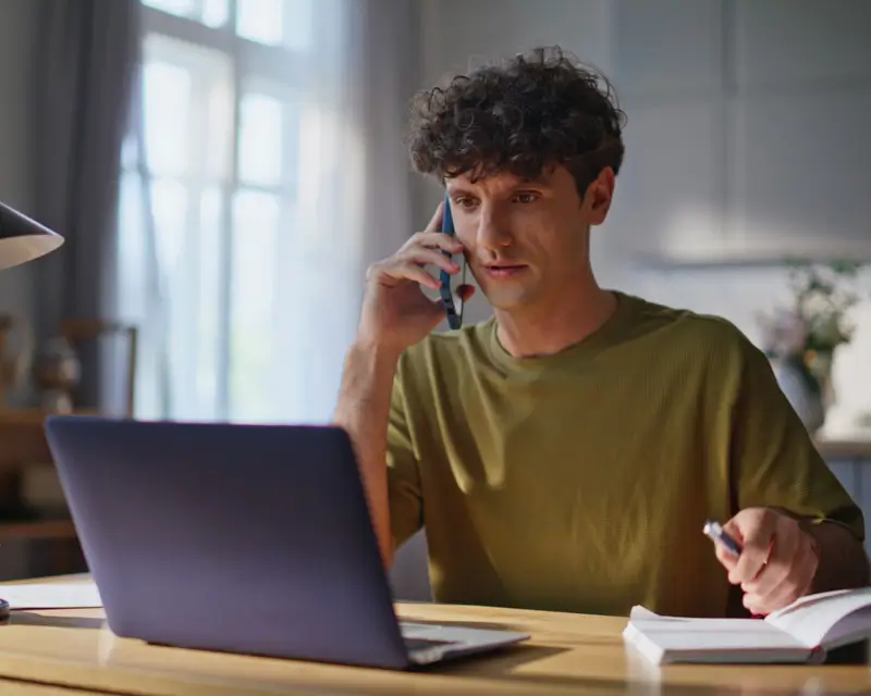 A young man with curly hair in an olive green t-shirt sits at a home desk talking on a mobile phone while holding a pen over an open notebook. A laptop is open in front of him, suggesting he is conducting a recruiting outreach or candidate screening call while taking notes.