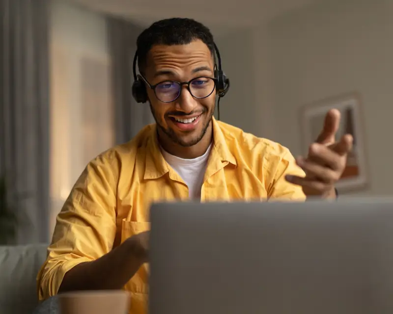 A smiling young man wearing glasses, a yellow shirt, and a professional headset gestures enthusiastically while looking at a laptop screen in a softly lit home environment. His engaged expression and headset suggest an active sales call or remote interview, reflecting the energy expected of high-performing sales candidates.