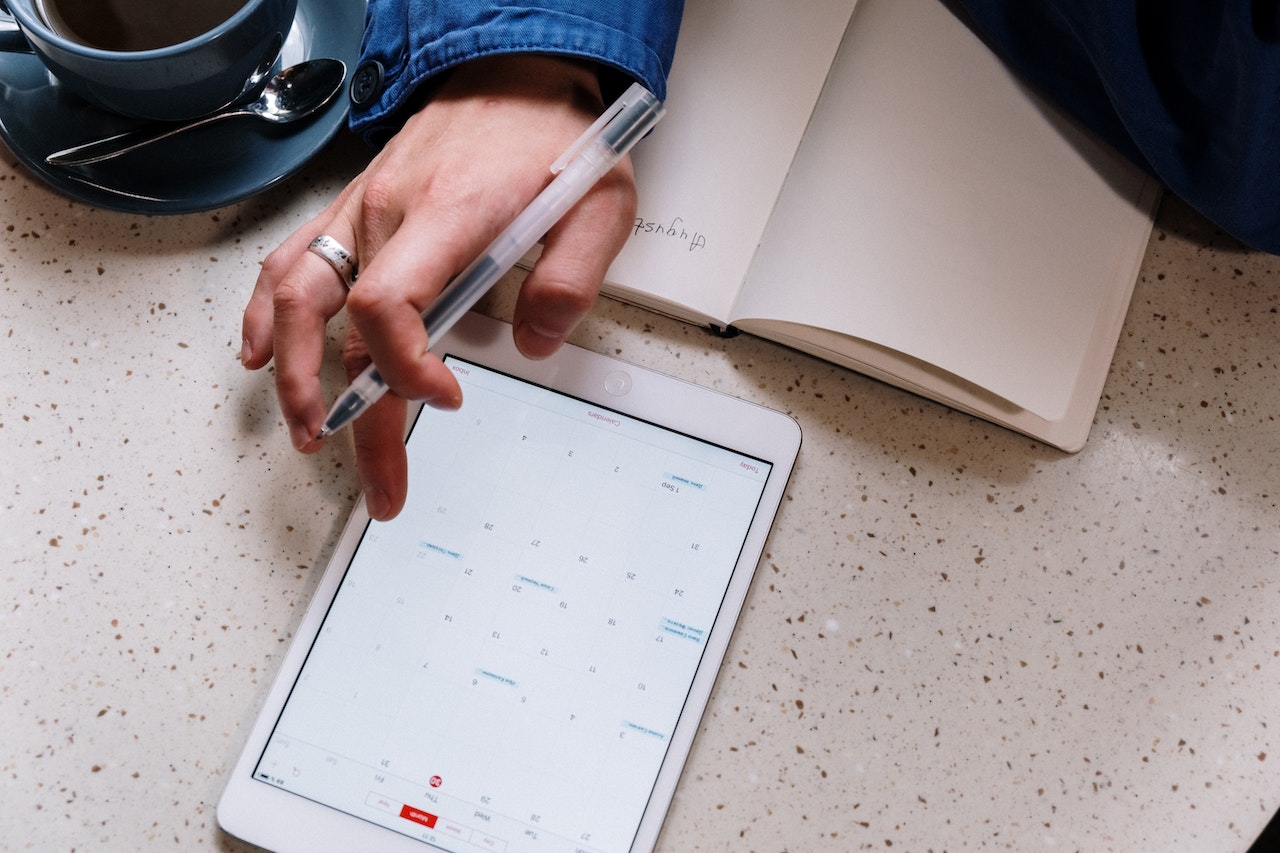 An overhead view of a hand using a stylus to interact with a calendar app on a white tablet, with a blank notebook and a cup of coffee also on the table. The image represents scheduling and time management, key elements of effective task delegation.