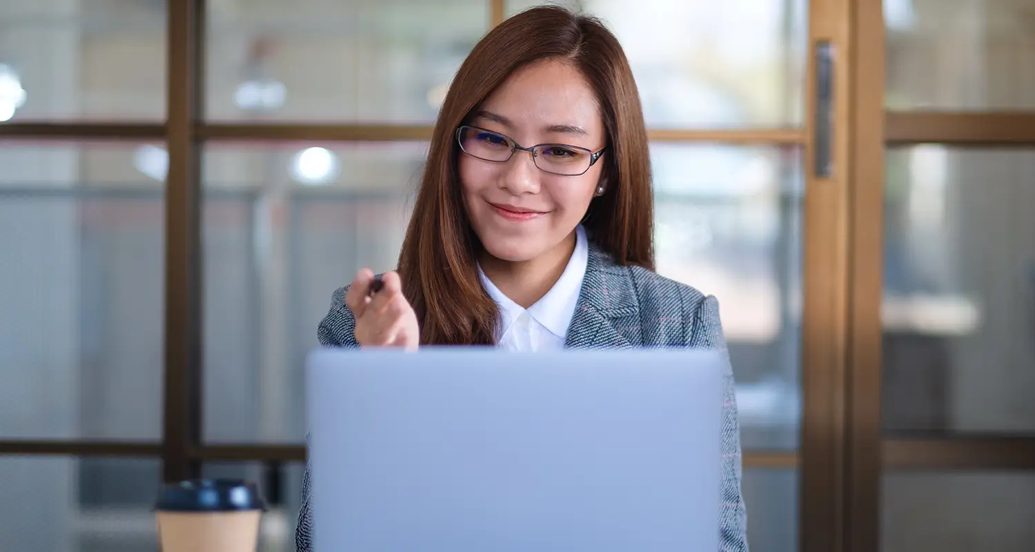 Smiling Asian woman with glasses holding a pen, seated at a laptop in a bright office, appearing confident and engaged in her work.