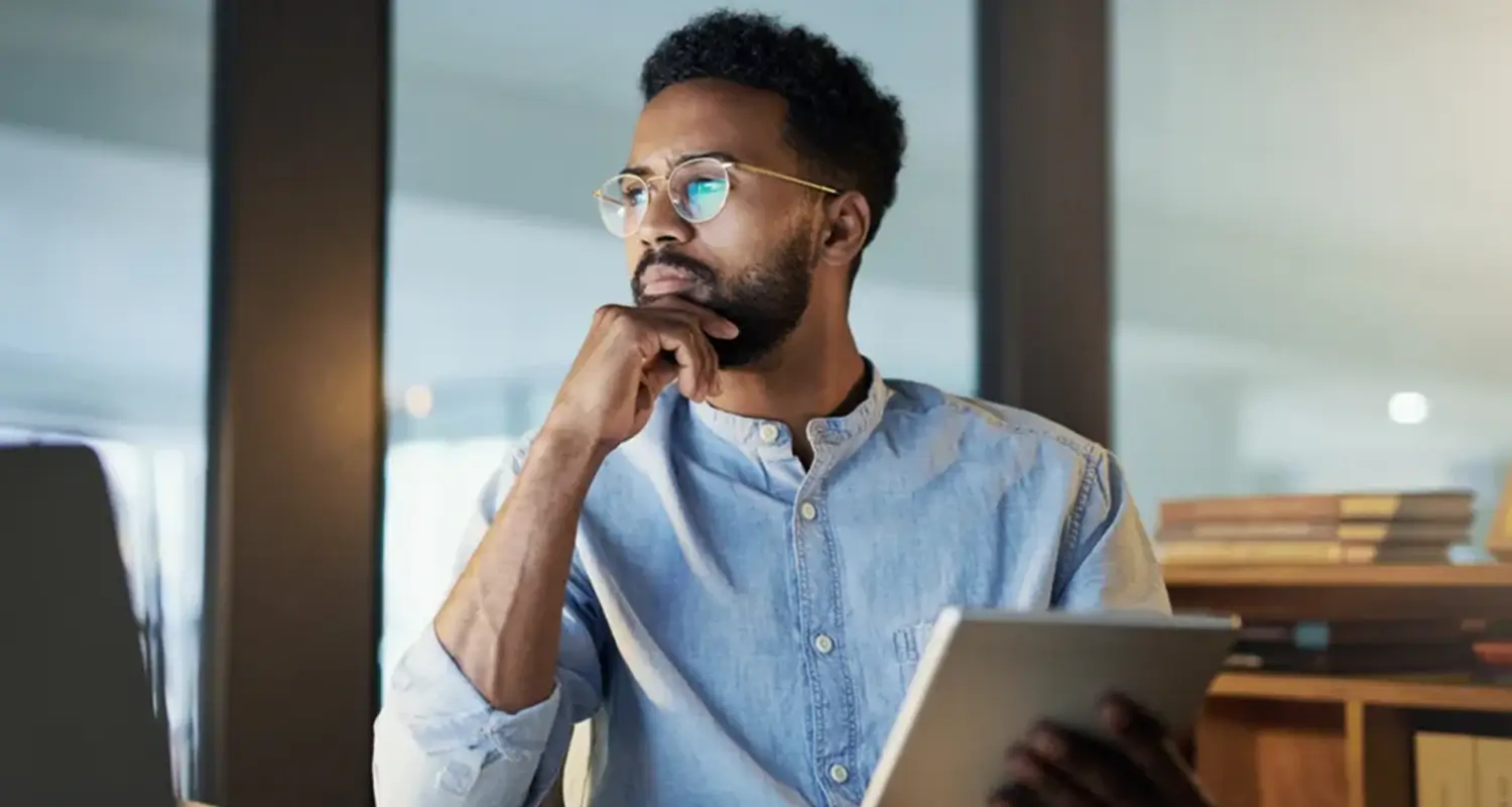Thoughtful Black professional man with glasses holding a tablet, looking upward while seated in a modern office environment.