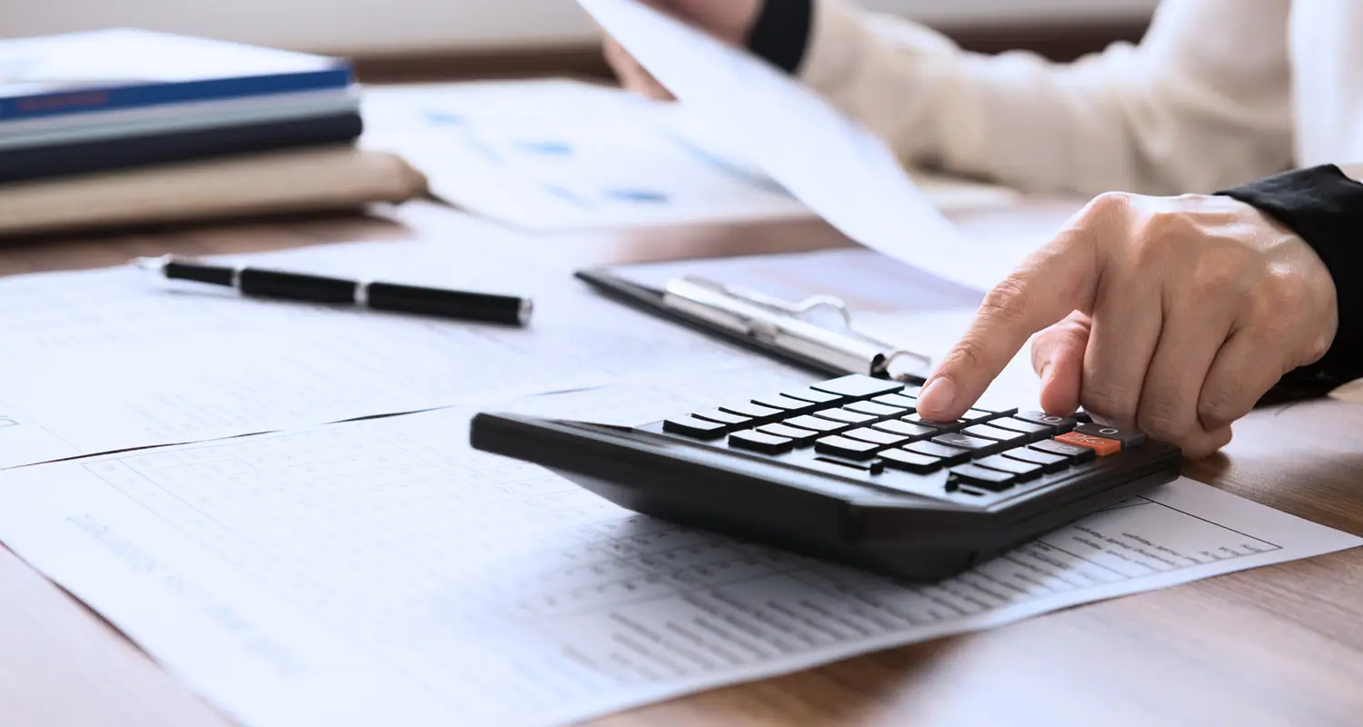 A close-up of hands operating a black desk calculator on top of printed financial documents, with a pen and notebook visible nearby. The scene depicts the core work of a bookkeeper managing financial records and calculations, representing the cost-saving value offshore bookkeepers bring to small and mid-sized businesses.