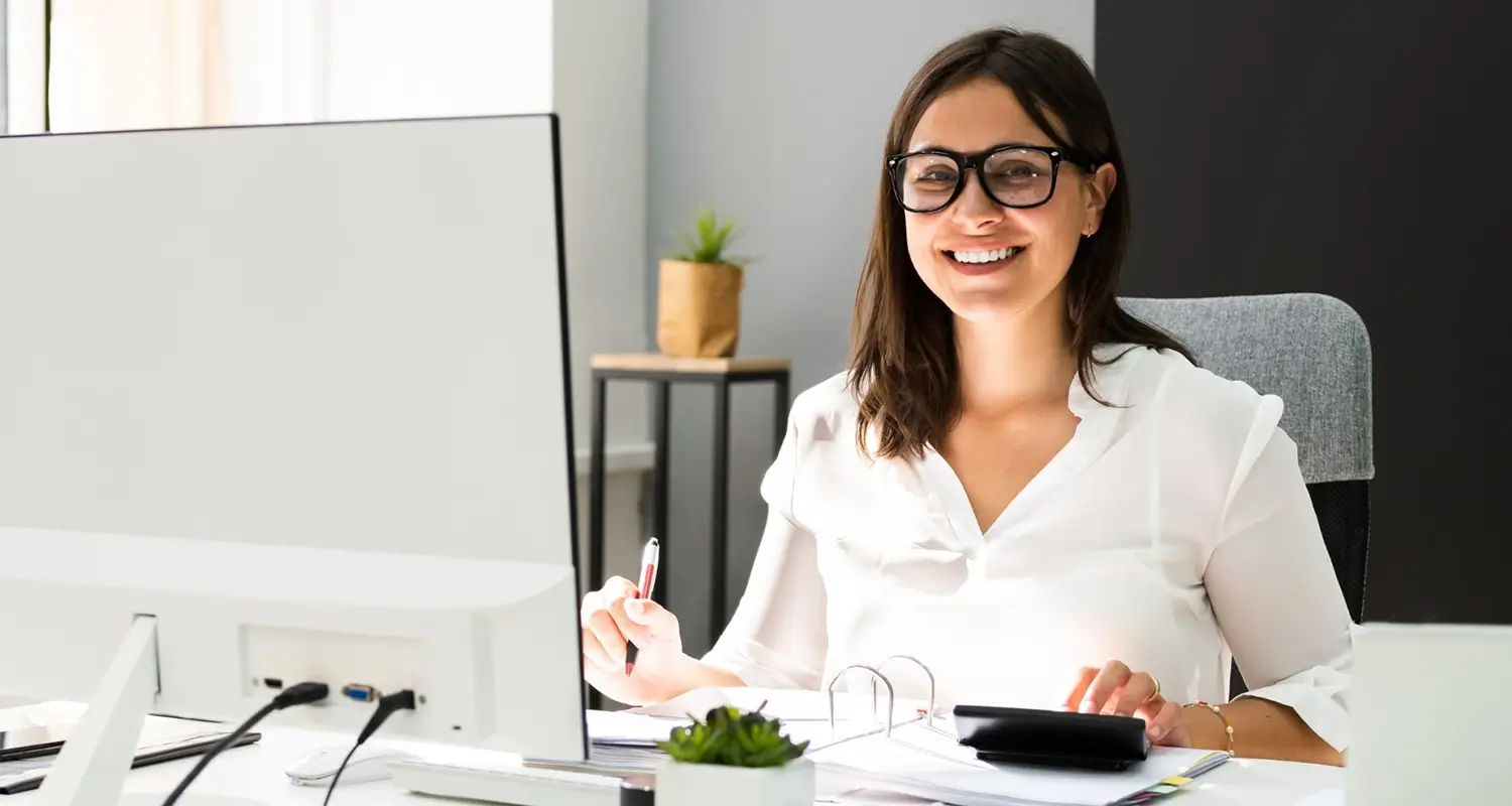 A smiling young woman with dark hair and glasses sits at a desk in a modern office, holding a pen beside a calculator and financial binder while facing a large computer monitor. Her professional and approachable appearance represents a skilled offshore bookkeeper handling financial records and accounts with accuracy and confidence.