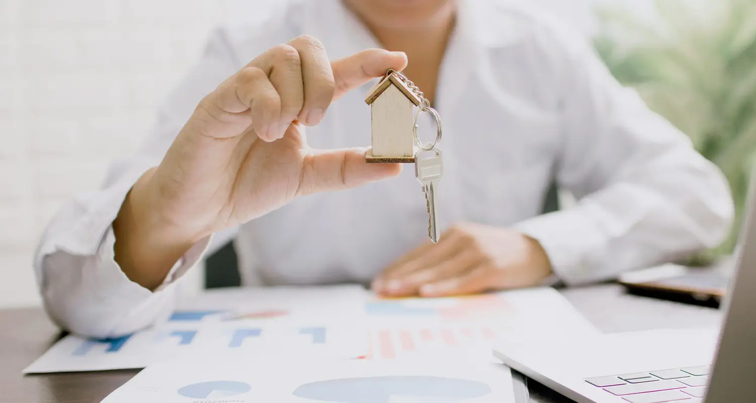 A person in a white shirt holds up a house-shaped keychain with two keys attached while sitting at a desk covered in real estate charts and a partially visible laptop. The image represents the property transaction and client management tasks that an offshore real estate virtual assistant handles on behalf of busy agents and brokers.