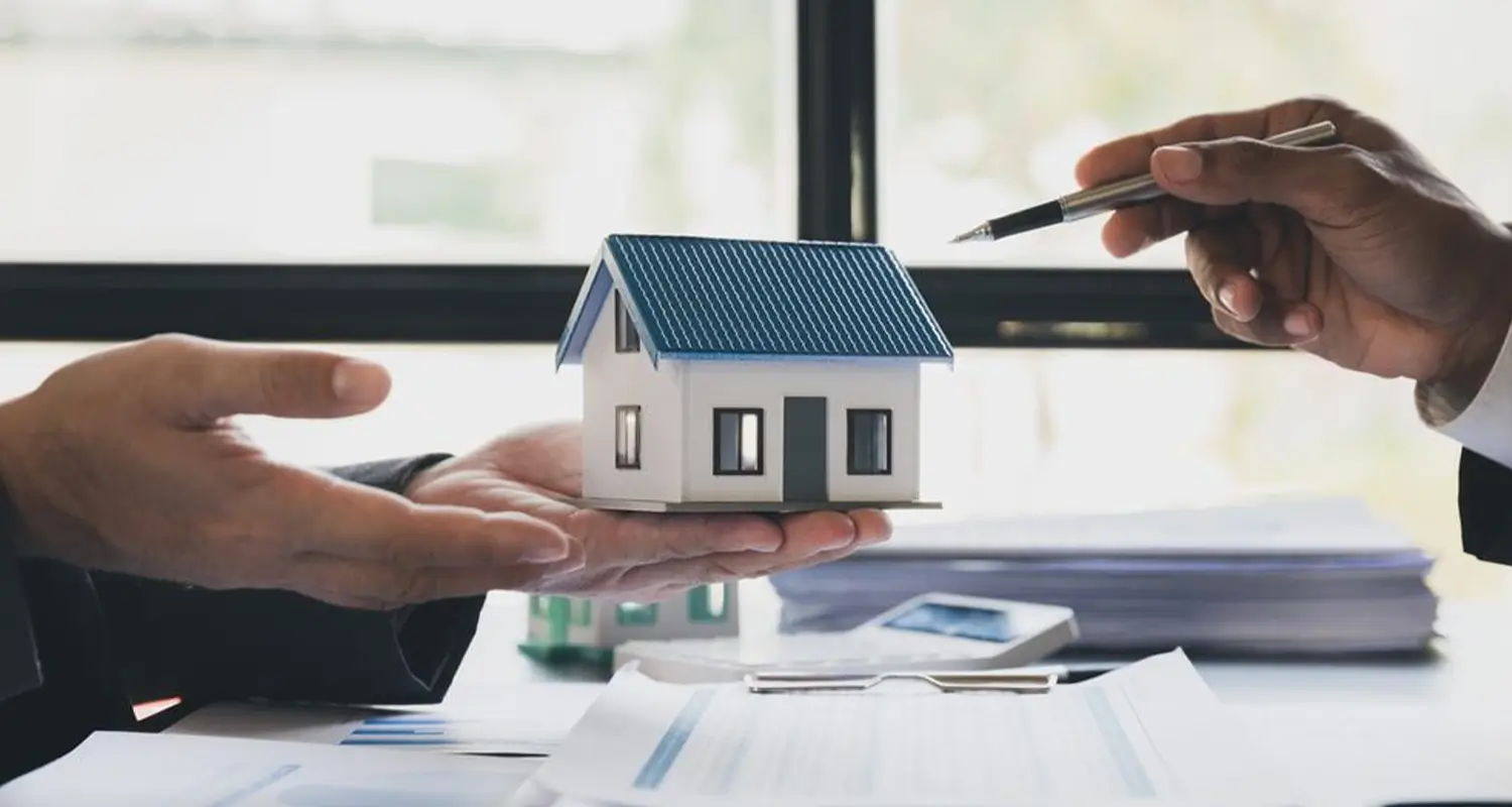 Two people in business attire face each other across a desk, with one holding a small model house while the other gestures with a pen toward documents. Property listings and financial paperwork are spread on the table, illustrating the type of transaction coordination and client support an offshore real estate virtual assistant provides.
