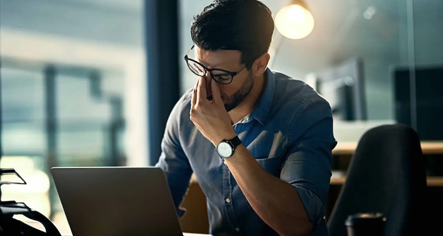 Stressed man in glasses rubbing his eyes while working late on a laptop in a dark office