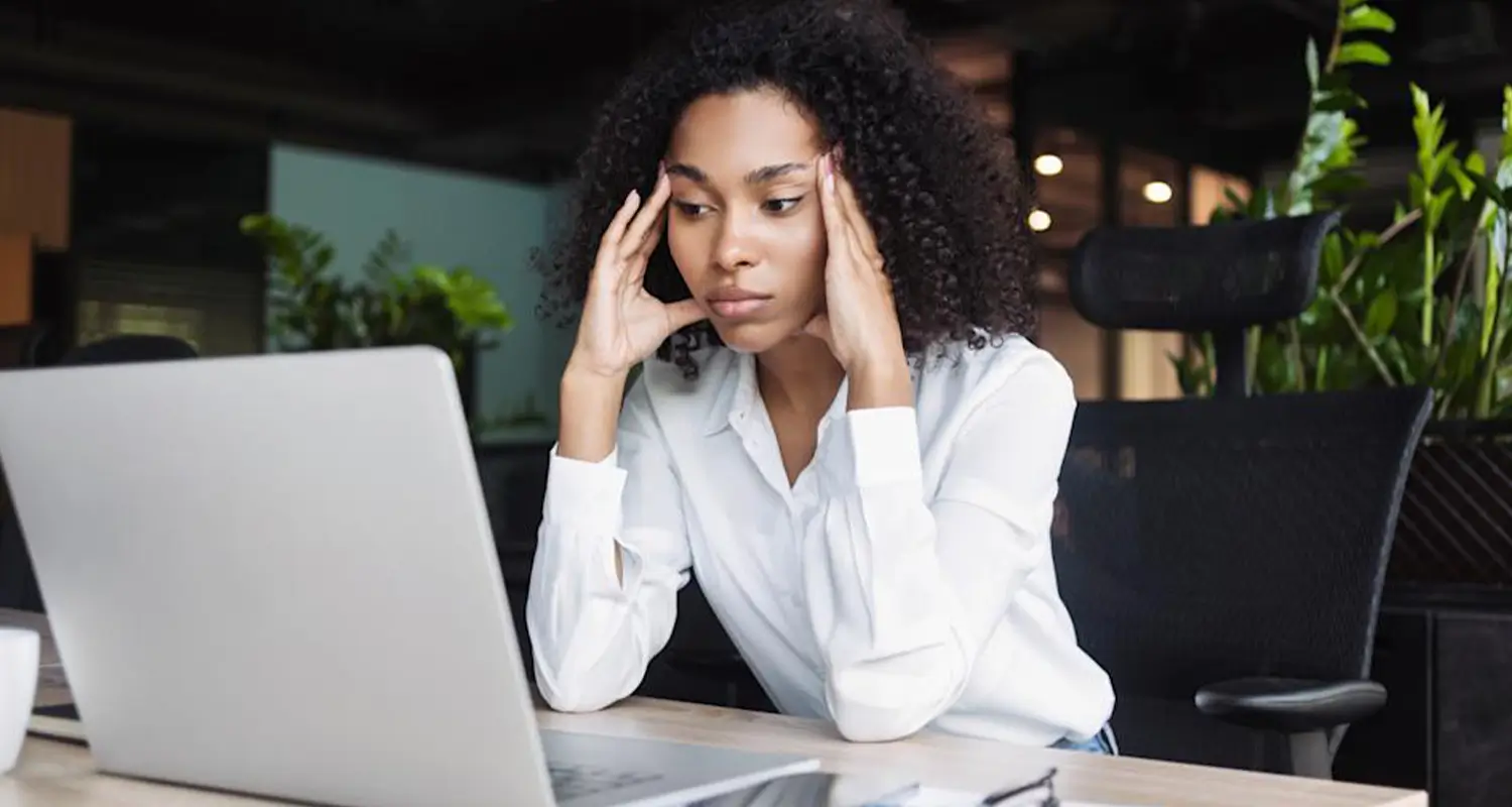 Stressed woman holding her temples while staring at a laptop screen in an open office