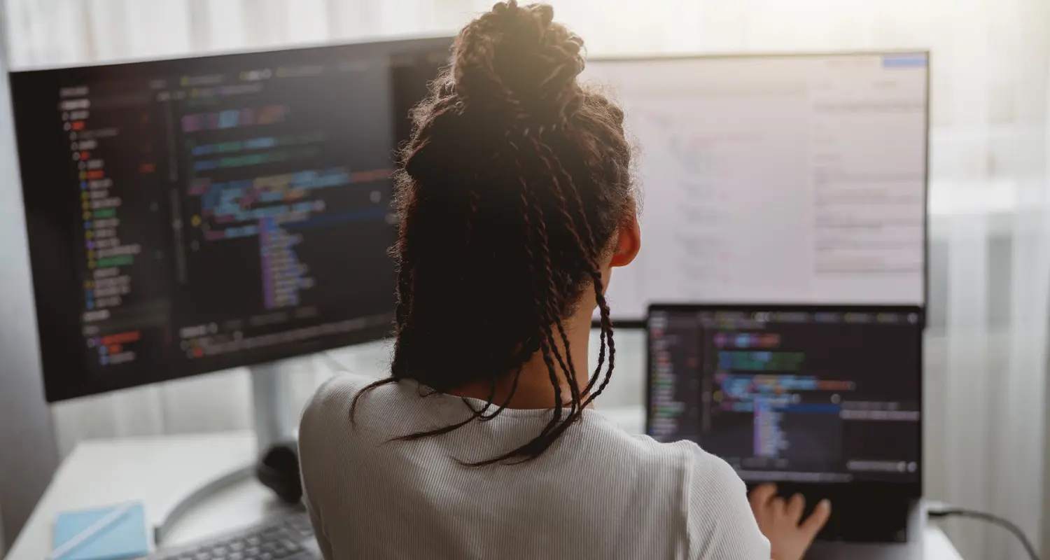 Female software engineer viewed from behind working at triple monitor setup with code