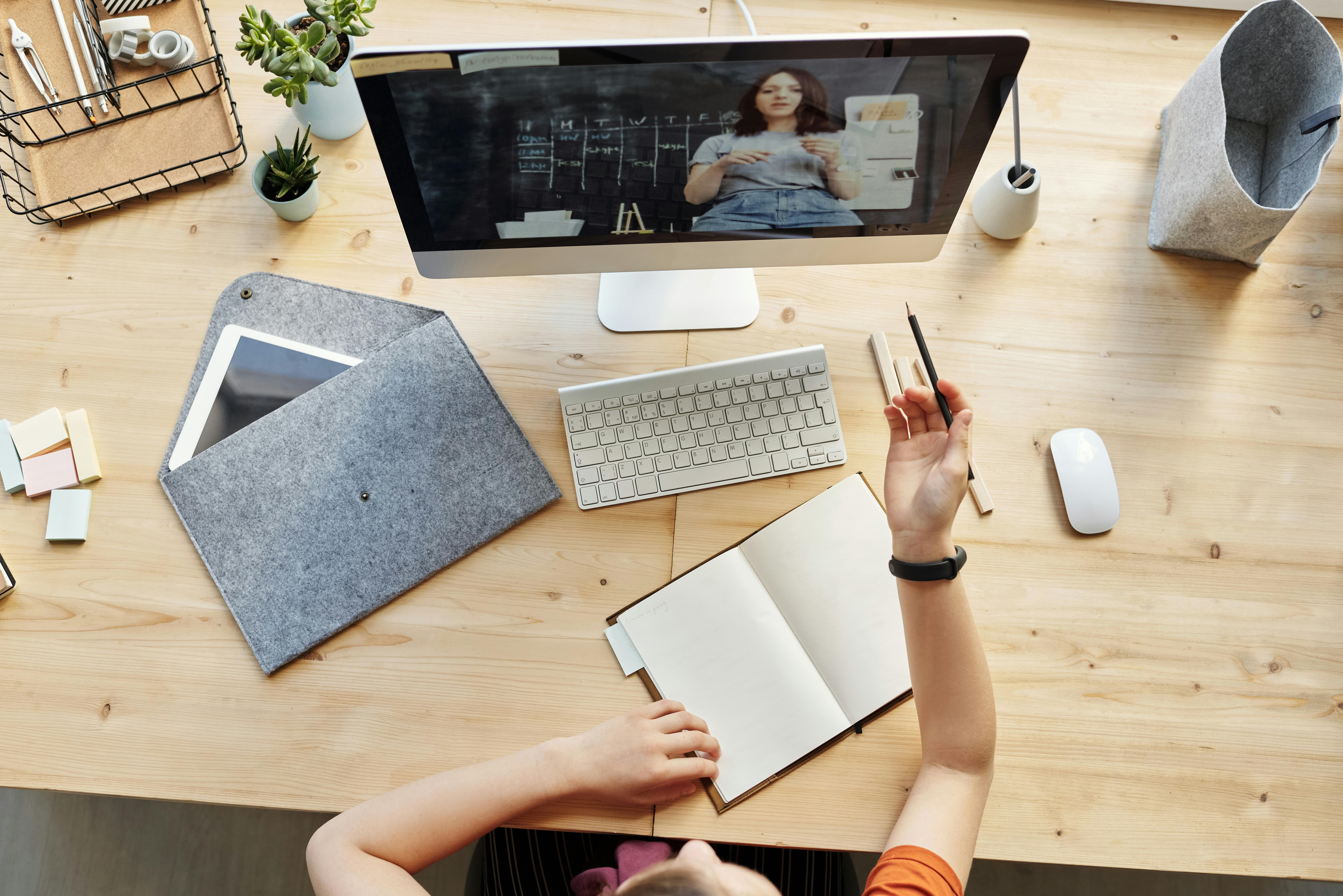 Overhead view of a person taking notes in a notebook at a desk with a desktop monitor showing a video call with a presenter