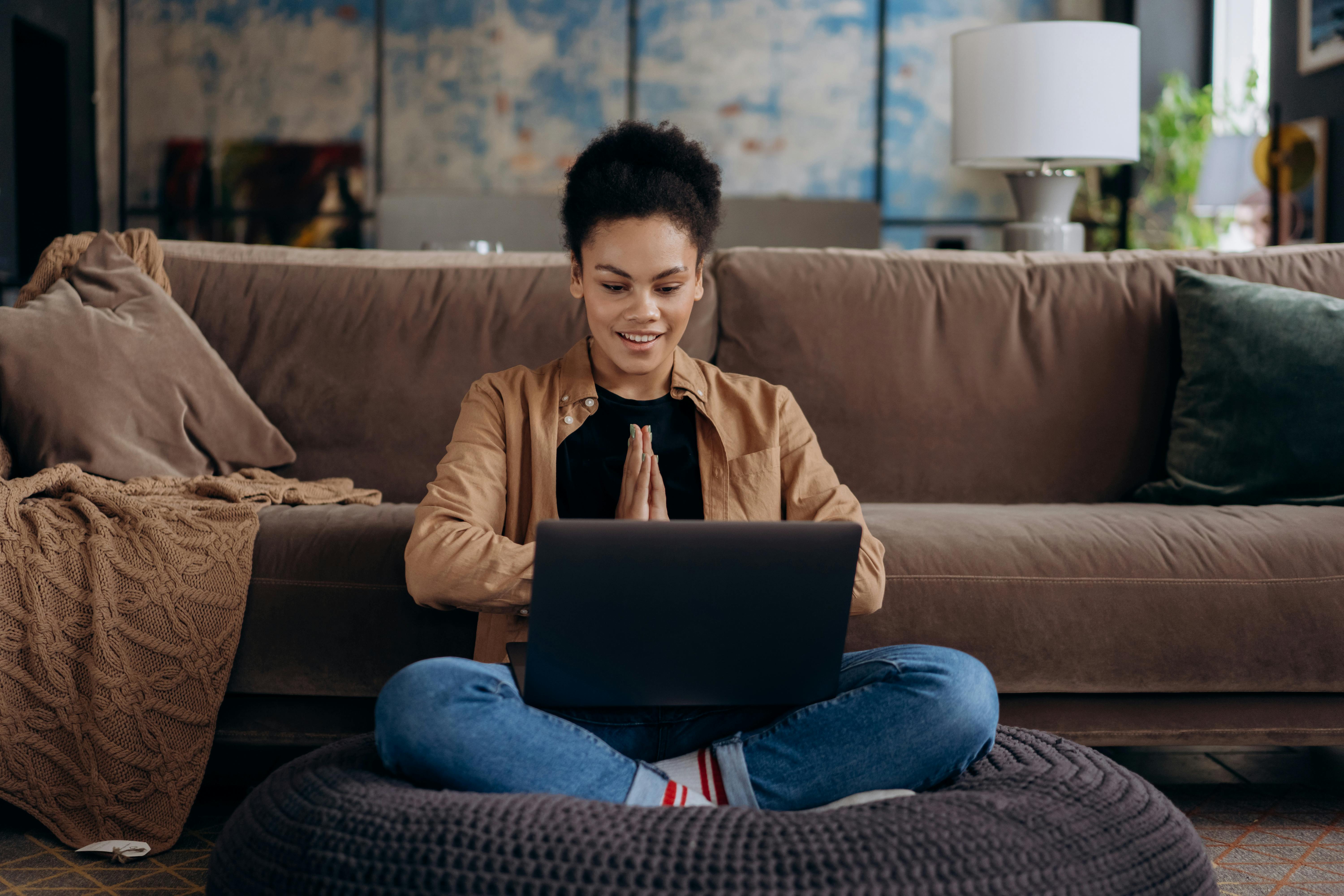 A young woman wearing a tan jacket sits cross-legged on a floor cushion in front of a couch, smiling while on a video call on her laptop. The cozy living room setting suggests she is a remote professional connecting with a client or employer from home.