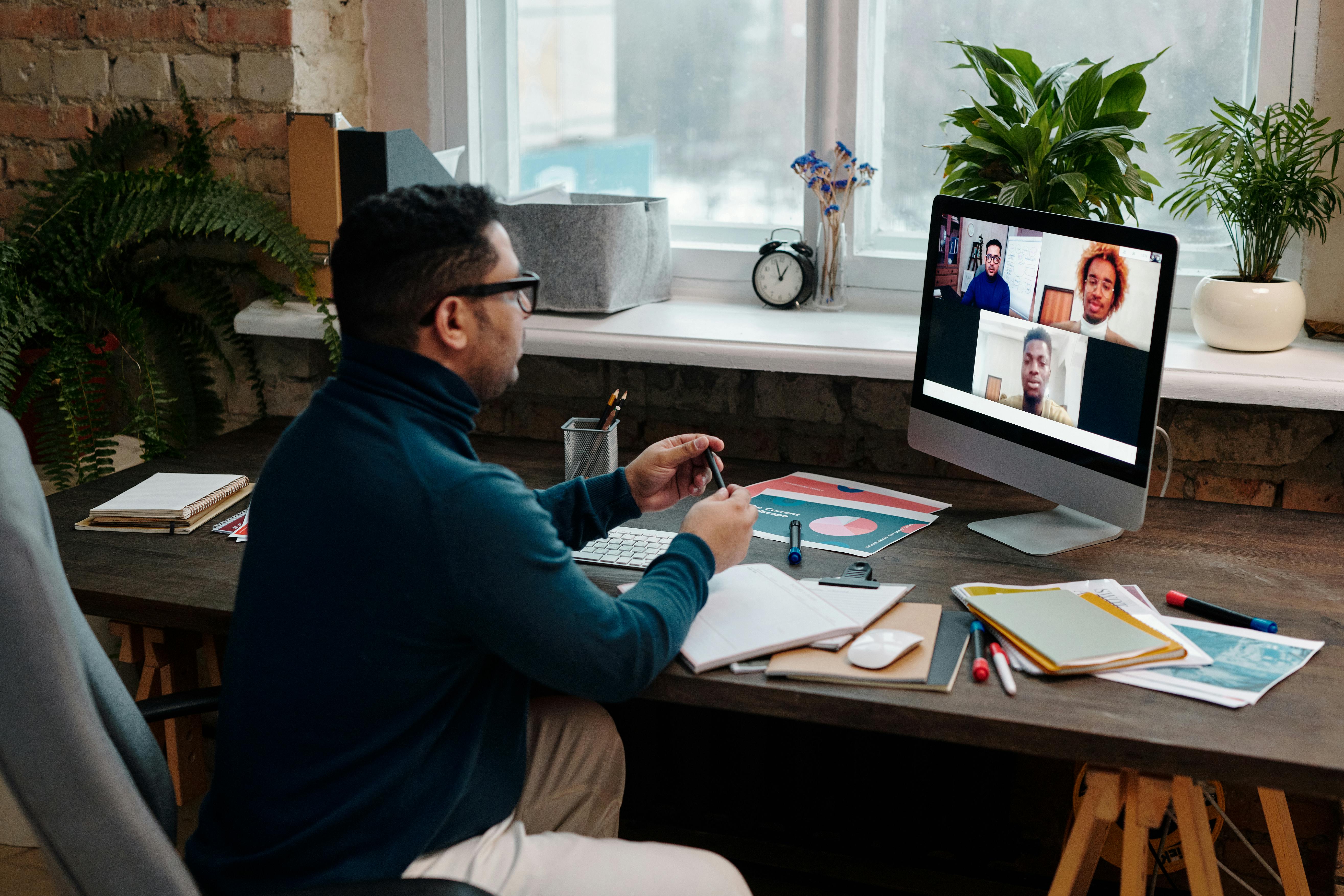 A man in glasses wearing a teal turtleneck sweater sits at a home office desk, participating in a video conference on a desktop monitor. The screen shows three colleagues in separate video tiles. The desk is organized with notebooks, markers, and a pie chart printout, surrounded by houseplants.