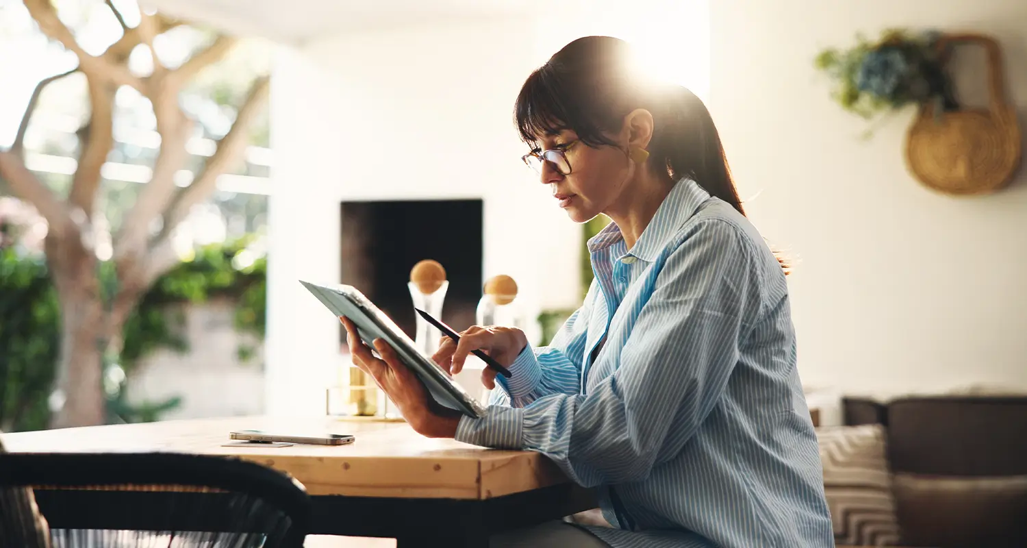 A woman in a blue striped shirt and glasses sits at a sunlit table in a bright living space, writing on a tablet with a stylus. Her phone rests nearby and warm natural light filters through surrounding greenery, depicting the flexibility and focus of a remote strategist working independently on global hiring plans.