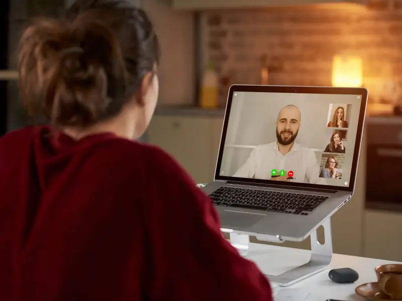 A woman with her hair up, wearing a red sweater, sits at a desk facing a laptop on a stand during a video call. The screen shows a bald man in a white shirt as the main speaker alongside two other participants in smaller panels, depicting a remote panel interview or hiring meeting.